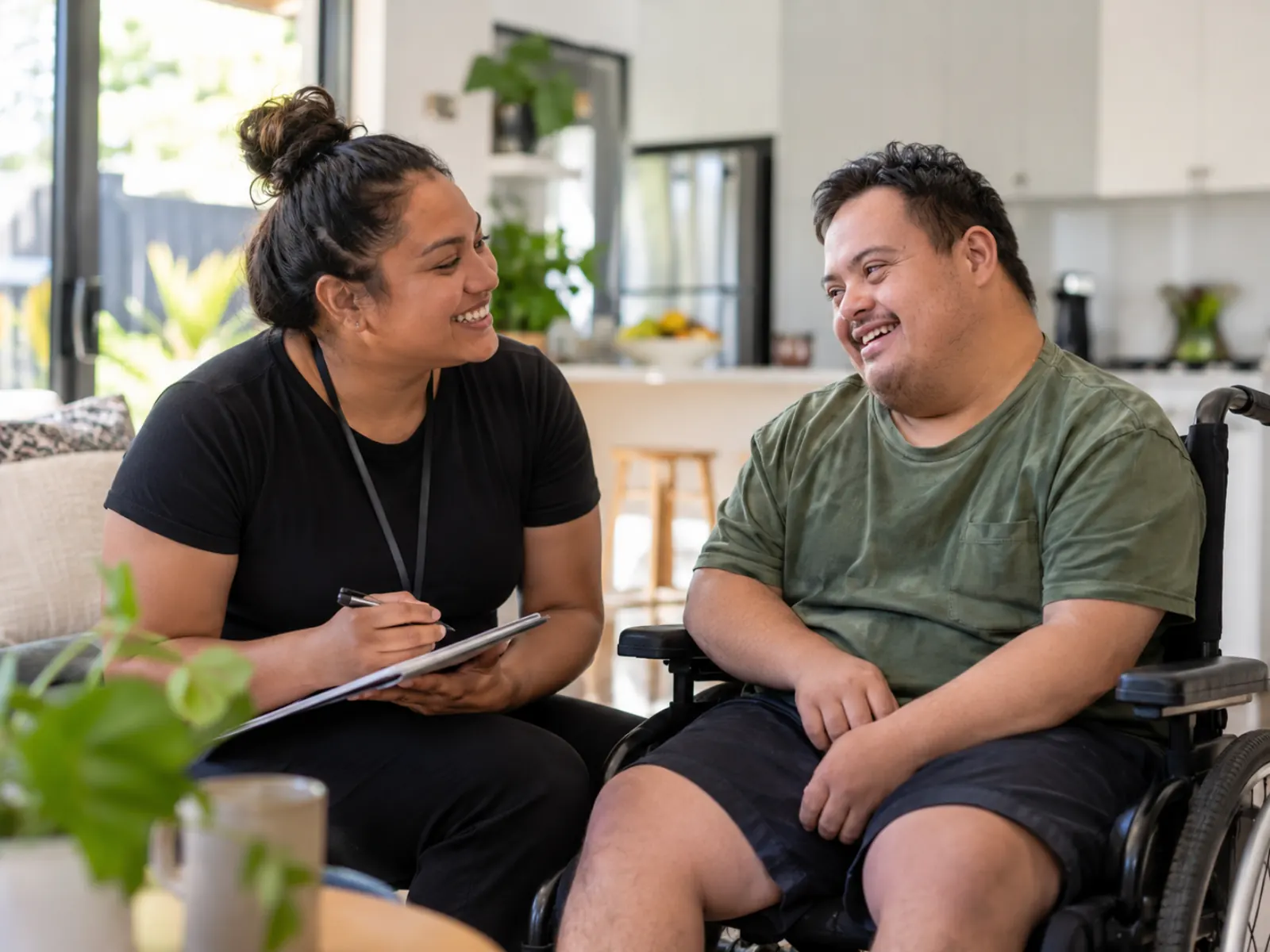 Support worker taking notes on a clipboard while chatting with a smiling young man in a wheelchair in a bright living room