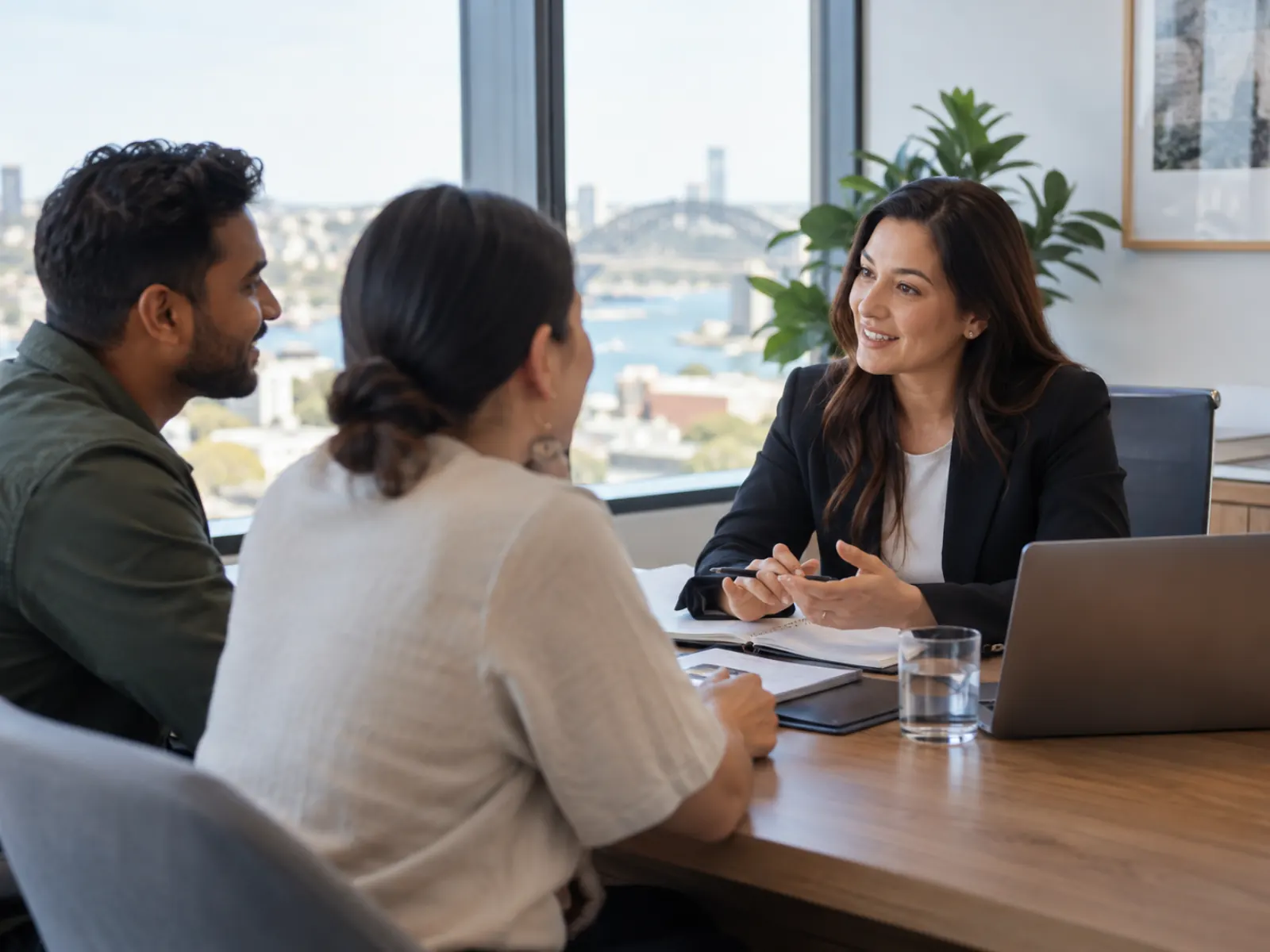 Adviser in a blazer talking with two clients across a meeting table with notes and a laptop, harbour view through the office window